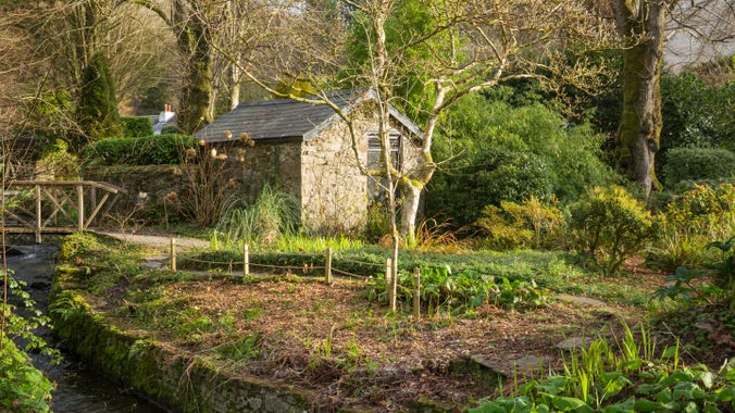 A small stone building in a garden with autumn leaves on the ground and trees in the background